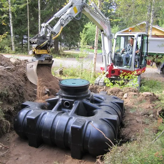 En grävmaskin från Cipax fyller skickligt smuts runt en stor svart septiktank av plast som delvis är nedgrävd i marken. En skicklig arbetare använder maskineriet med precision, placerat utomhus nära träd och en slingrande grusväg.