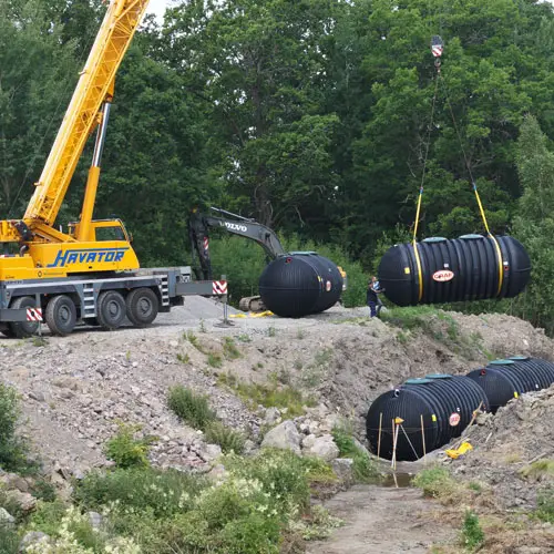 En kran lyfter ett stort Conclean Minireningsverk cylindriskt föremål upp på en byggarbetsplats medan en person i närheten observerar. Ett annat liknande föremål ligger på marken. Träd och stenig terräng omger området, vilket bidrar till den robusta bakgrunden.
