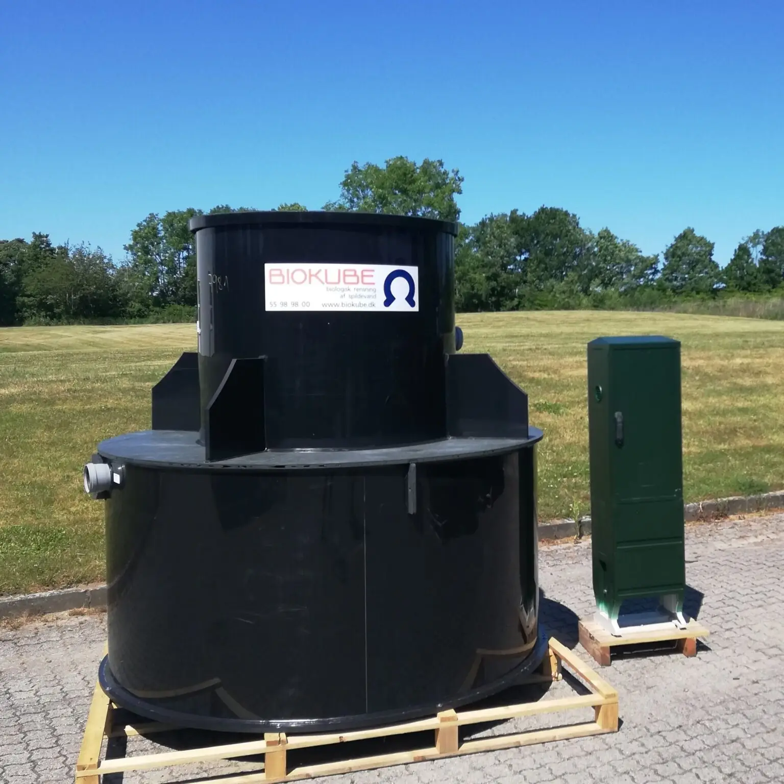 En stor svart cylindrisk Biokube-enhet, en del av ett Minireningsverk-system, placeras utomhus på en pall. Den har en liten etikett med logotyp och kontaktinformation. Bredvid den står ett högt, grönt kontrollskåp. Båda vilar på en asfalterad yta, inramad av gräs och träd i bakgrunden.