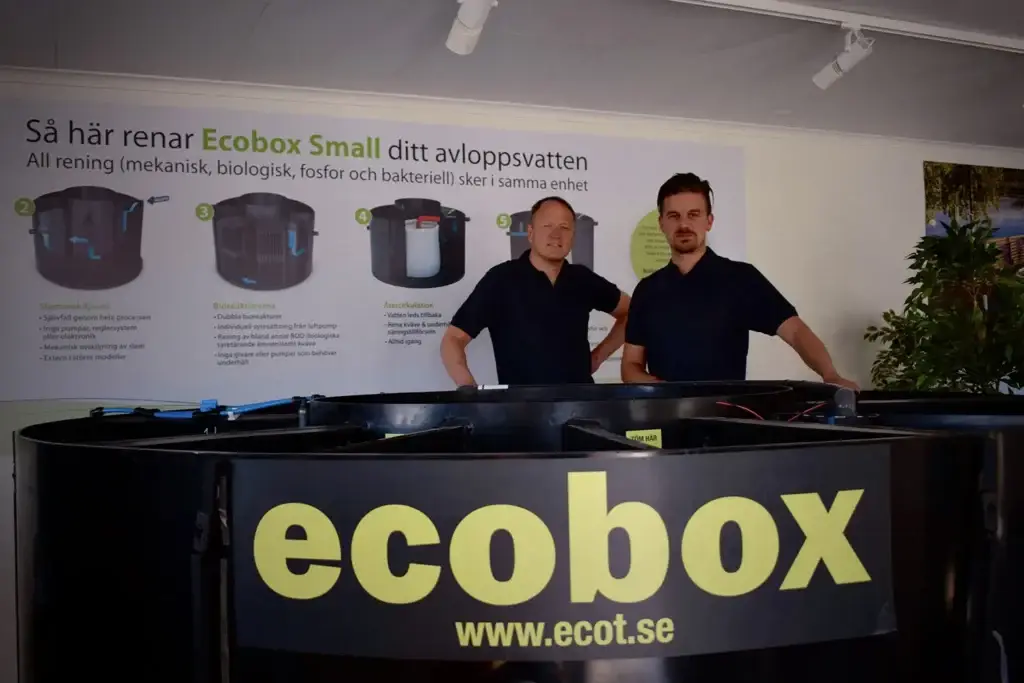 Två personer står bakom en stor svart tank märkt "ecobox" med en webbadress. En skylt bakom dem förklarar hur Ecobox Small effektivt renar avloppsvatten, med diagram och text på svenska. Frodiga växter flankerar eftertänksamt den informativa displayen.