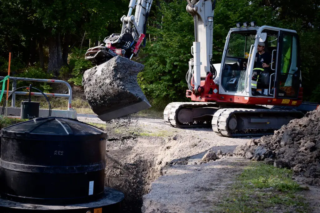 En byggnadsarbetare kör en liten grävmaskin och använder sin skopa för att hälla grus i ett stort cylindriskt hål för en smarta avloppslösning med minireningsverk. Platsen är omgiven av träd och området verkar vara en del av en väg eller stig som är under uppbyggnad.