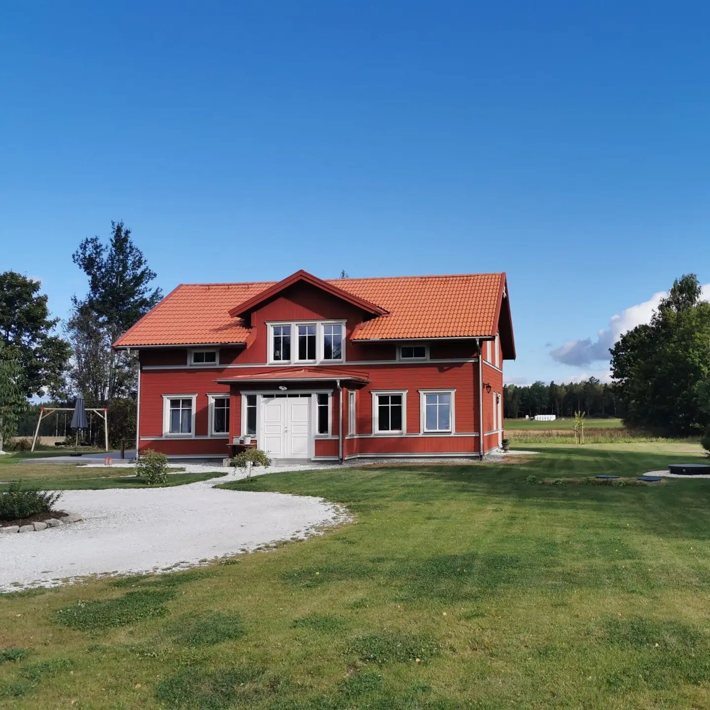 Ett rött hus i två våningar med vit dörr och vita fönsterkarmar står på en stor grön gräsmatta under en klarblå himmel. Träd kantar bakgrunden och en svängd grusad uppfart leder förbi ett diskret Biokube-system på framsidan.