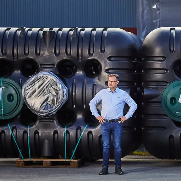 En man med glasögon och ljusblå skjorta står med händerna på höfterna framför två stora svarta Cipax industritankar med gröna runda öppningar, placerade utomhus på en lastpall.