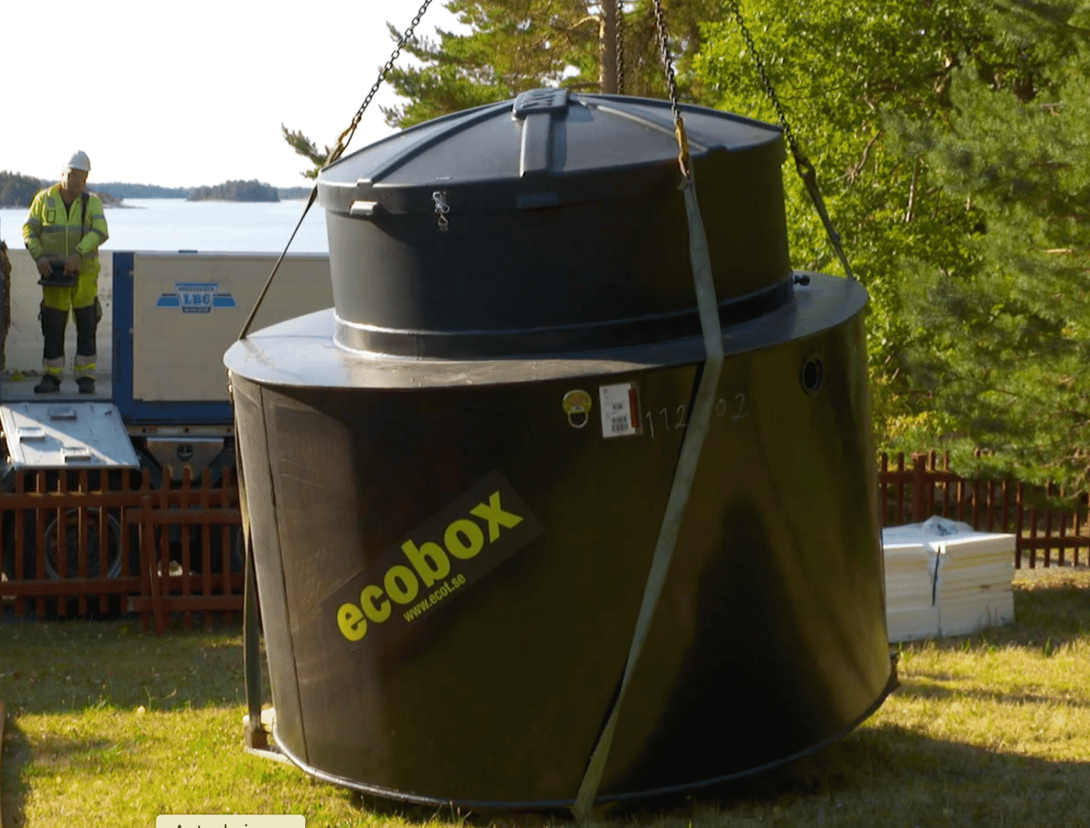 En stor svart cylinderformad Ecobox-behållare lyfts med kranremmar utomhus, med en arbetare i skyddsutrustning och ett skogslandskap i bakgrunden.