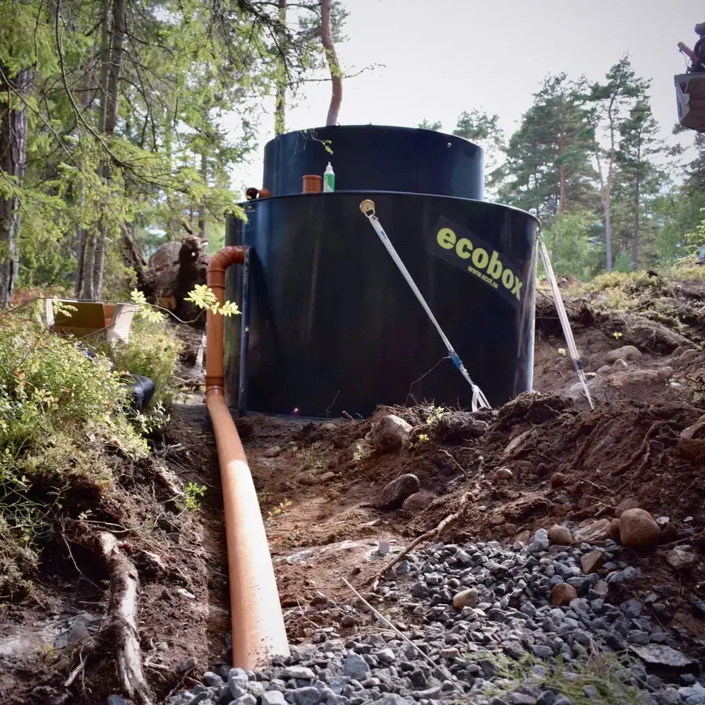 En stor svart cylindrisk minireningsverk märkt "ecobox" är installerad utomhus i ett skogsområde, med ett orange rör anslutet till den och viss utrustning placerad ovanpå.