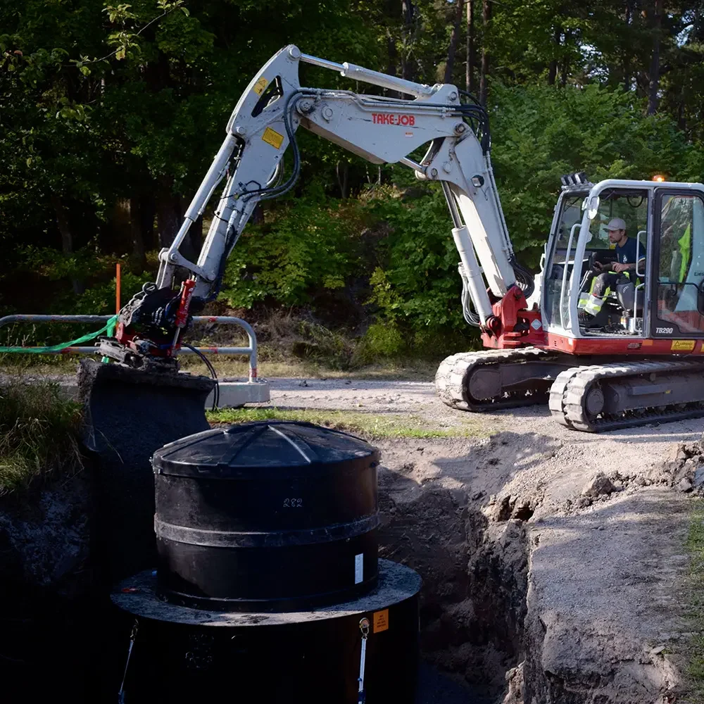 En person kör en kompakt grävmaskin intill en byggarbetsplats med en stor svart cylindrisk tank som delvis är nedgrävd i marken. I bakgrunden syns jordhögar och träd.