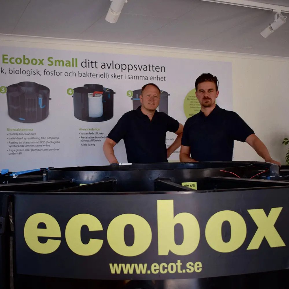 Två män står bakom en stor svart behållare märkt "ecobox" med webbplatsen ecot.se. I bakgrunden syns en affisch som beskriver Ecobox Small, ett minireningsverk, på svenska.